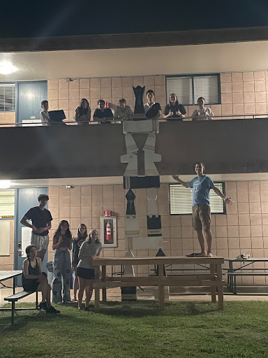 A tower of giant wooden chess pieces as tall as a second-story balcony. Students are standing in the dark on the balcony as well as around the base of the stack showing off their work.