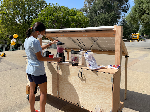  A woman stands in front of a blender connected to a cart located outdoors