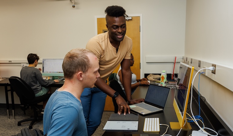 Student in blue shirt and mentor in peach shirt discussing results on shared computer screen at a long brown table