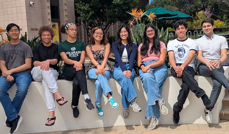 Professor Brucks with his seven spring 2026 researchers sitting outside on a low white wall with legs crossed in the same direction