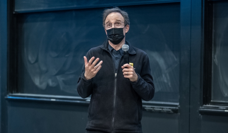 Masked professor stands in front of chalkboard holding microphone