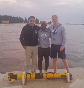 Timmy Gambin (University of Malta), poses with Harvey Mudd College students Eric Contee ’19 and Russell Bingham ’20 with an autonomous vehicle at their feet