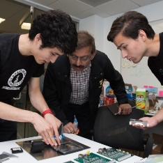 Two students and Prof Gokli look at circuit boards on table