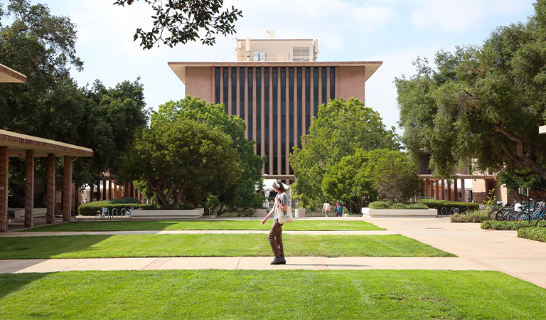 A student holding a tote bag and wearing headphones walks across campus.