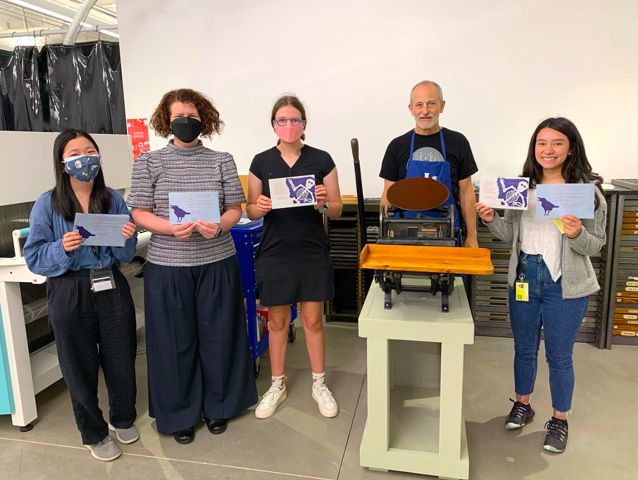 Three students and two adults standing behind the printing press, holding papers with an image and text that they made with the printing press.