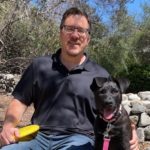 Jon Jacobsen sits on a rock next to dog Roxie Jacobsen, during a sunny day.