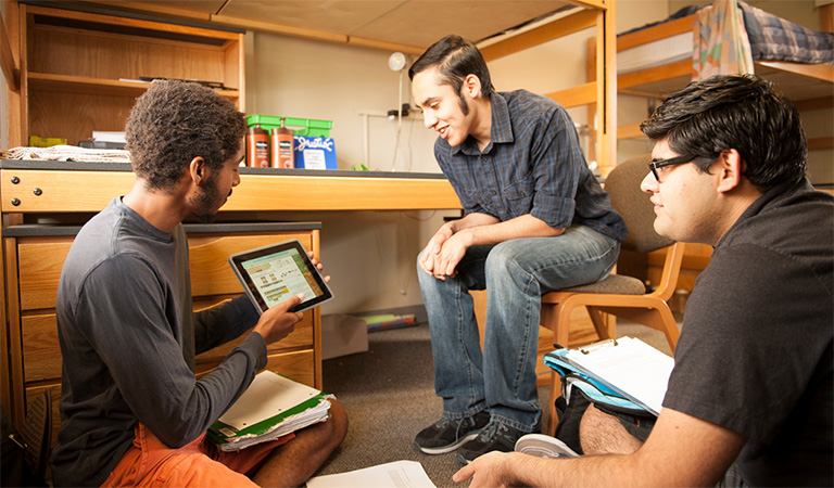 Three students in a dorm room studying on a tablet