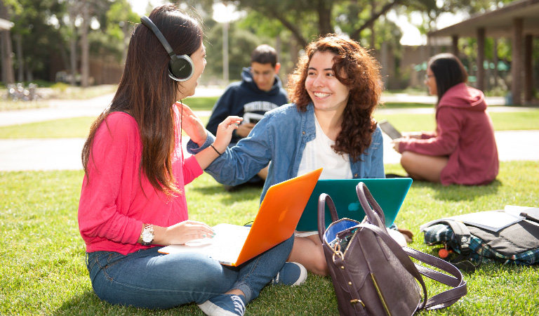 Two students laughing and sitting on a lawn with laptops in their laps.