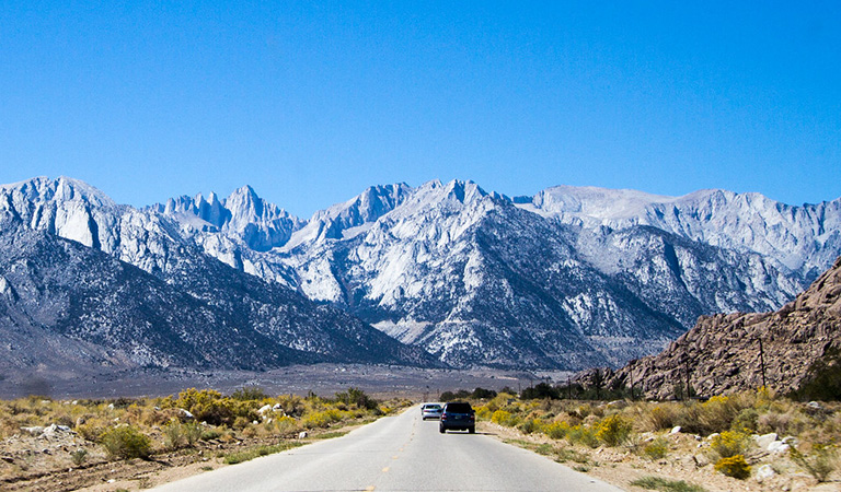 Cars driving down a road towards snow-covered mountains.